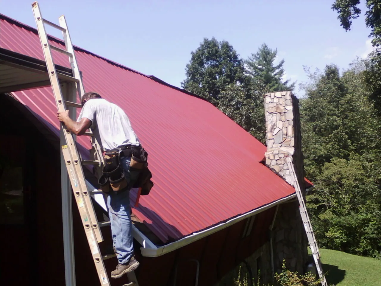 Roofer on a ladder beside a red metal roof and stone chimney — Soffit & Fascia Repair by Roofing Brothers in Helotes